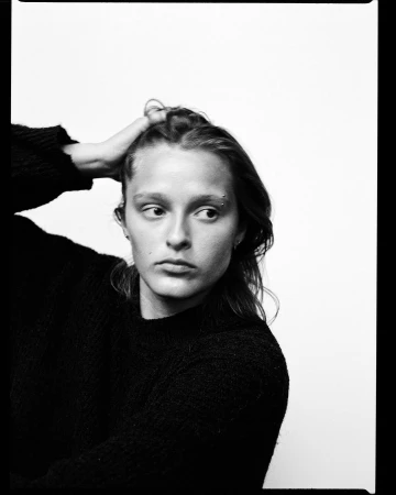 Monochrome portrait of a young adult woman with a hand in her hair, wearing a dark sweater, against a plain white backdrop.