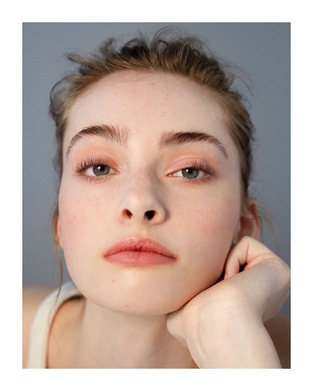 Close-up portrait of a young Caucasian woman with natural makeup, posing with hand on chin against a grey backdrop in a photo studio.