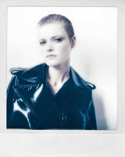 Close-up portrait of a fair-skinned woman with slicked-back hair wearing a dark, shiny jacket against a bright, diffused background.