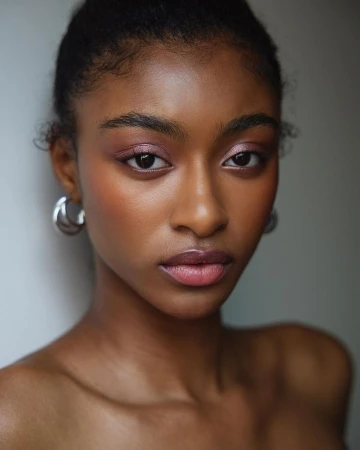 Close up portrait of a Black woman with subtle pink eyeshadow and silver hoop earrings against a neutral background.