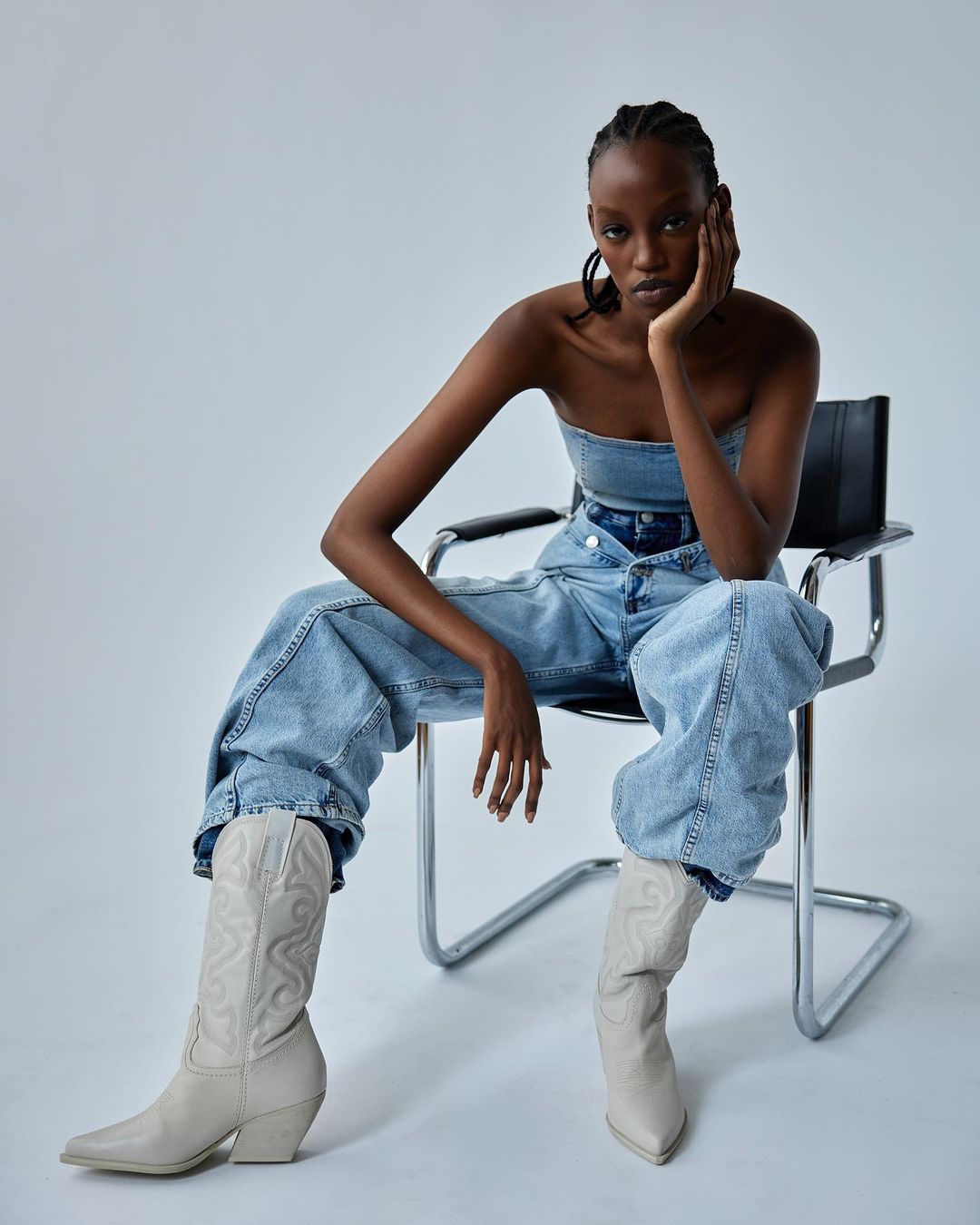 A Black female model sits on a chair wearing a denim outfit and boots in a light studio. The model poses with her hand to her face.