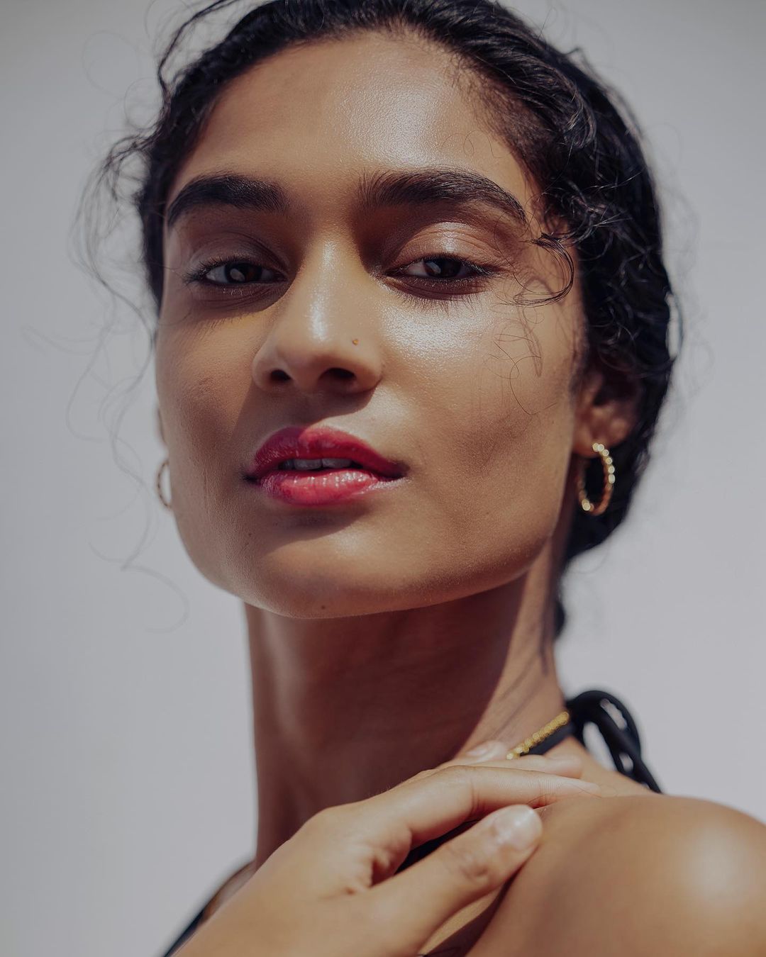 Close-up portrait of a woman with dark curly hair wearing gold hoop earrings and red lipstick against a white background