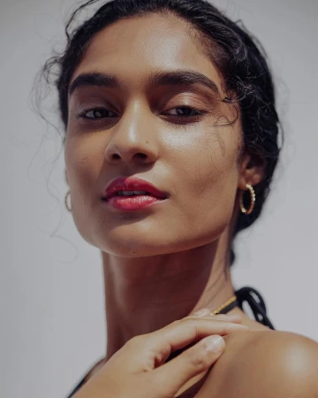 Close-up portrait of a woman with dark curly hair wearing gold hoop earrings and red lipstick against a white background