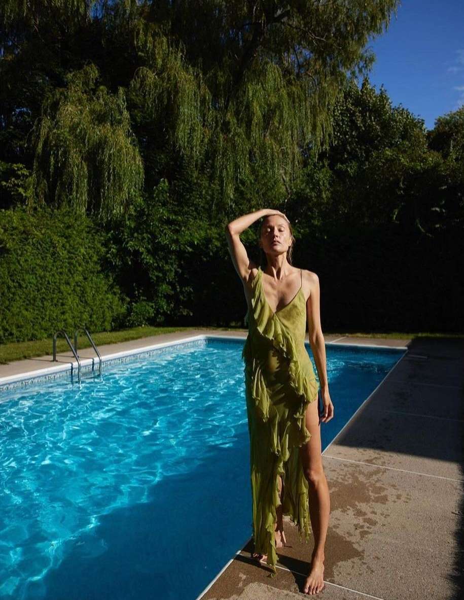 A slender woman in a flowing green dress poses elegantly near a bright blue swimming pool outdoors on a sunny day, with green foliage.