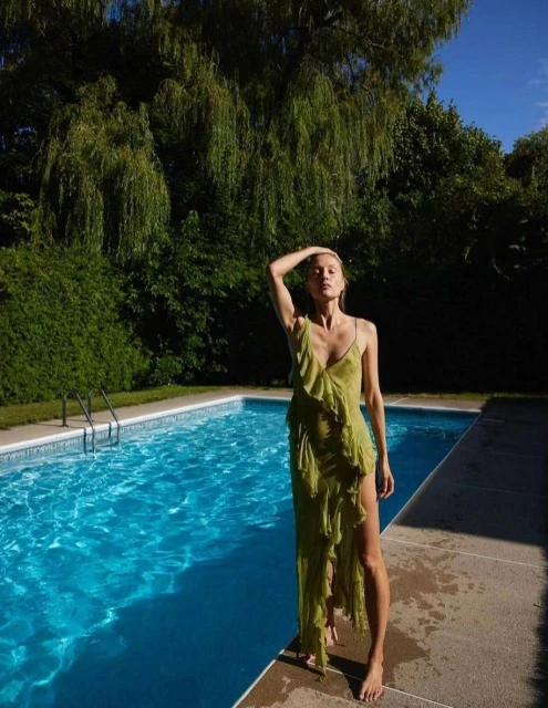 A slender woman in a flowing green dress poses elegantly near a bright blue swimming pool outdoors on a sunny day, with green foliage.