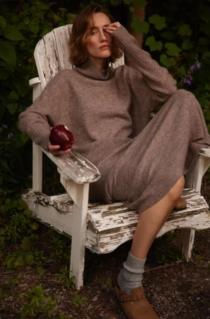 Woman in brown turtleneck sweater seated in a weathered white wooden chair holding a red apple outdoors