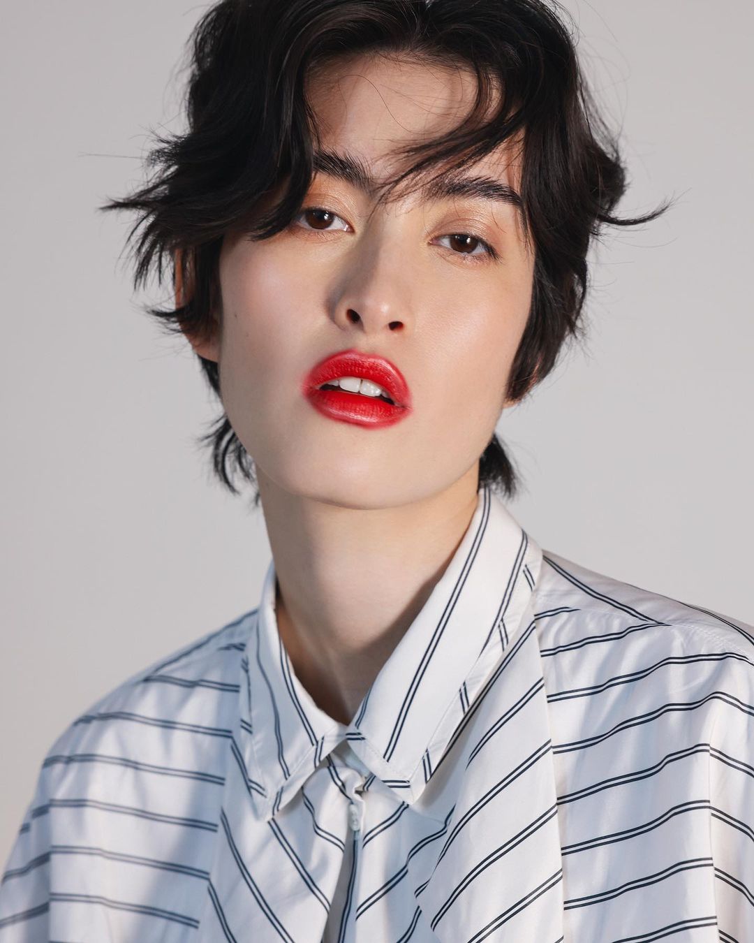 Close-up portrait of a stylish adult with dark hair, fair skin, and red lipstick posing in a striped shirt against a neutral background.