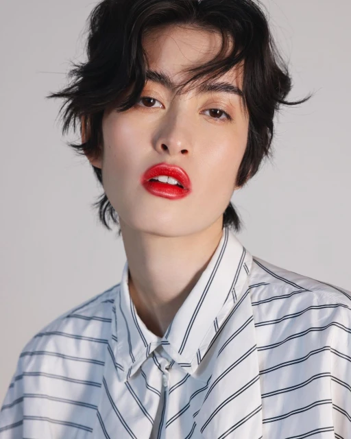 Close-up portrait of a stylish adult with dark hair, fair skin, and red lipstick posing in a striped shirt against a neutral background.