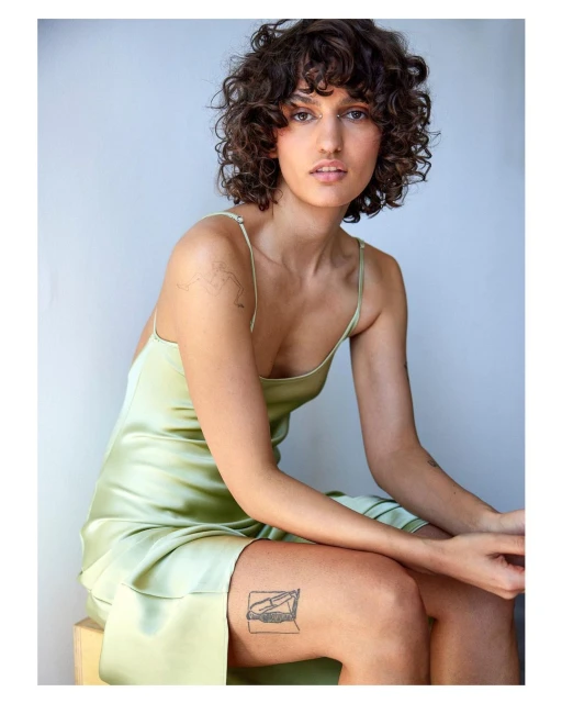 Woman with curly hair in a light green satin dress sitting on a wooden box in a studio with natural lighting, portrait shot.
