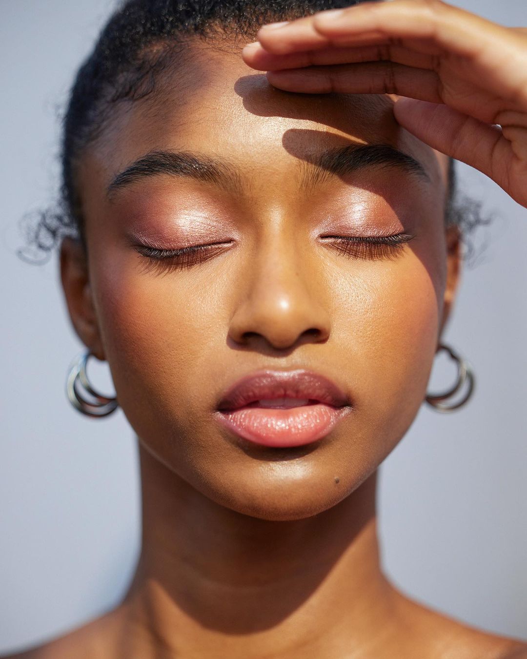 Close-up portrait of a dark-skinned Black woman shielding her eyes, with soft makeup, closed eyes, hoop earrings, and soft studio lighting.