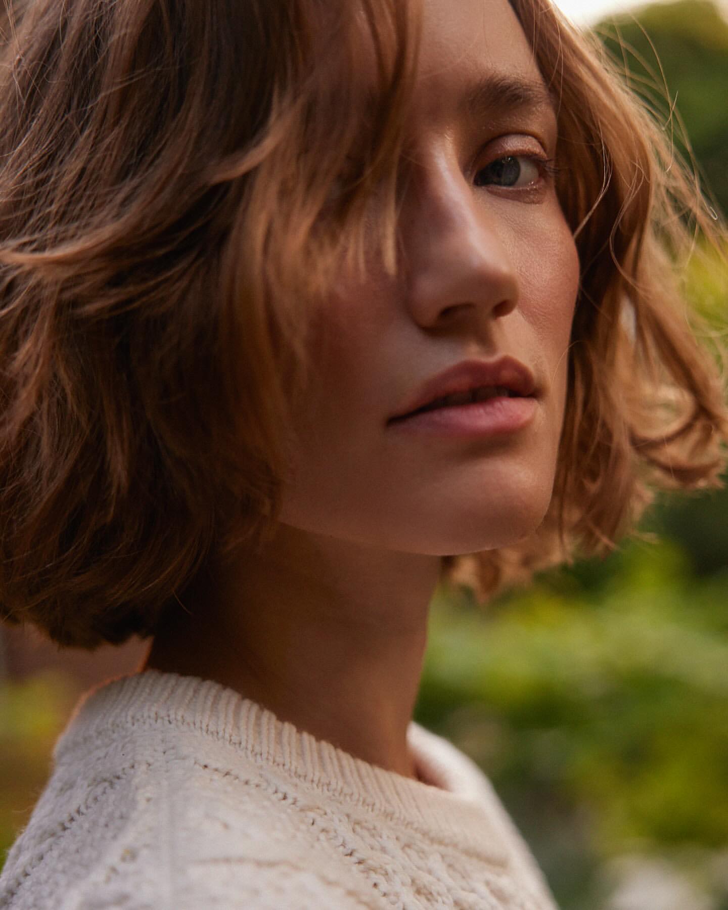 Close-up portrait of a young adult woman with short wavy hair and wearing a white patterned sweater against a blurred green background.
