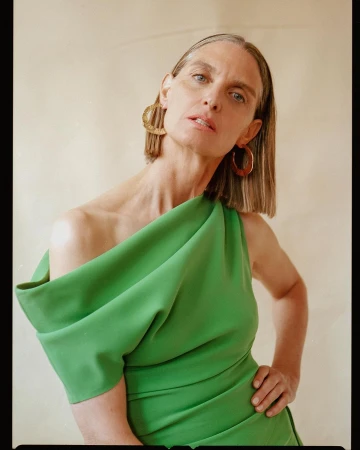 Portrait of an older woman with chin-length hair wearing a green one-shoulder dress and large hoop earrings against a neutral backdrop