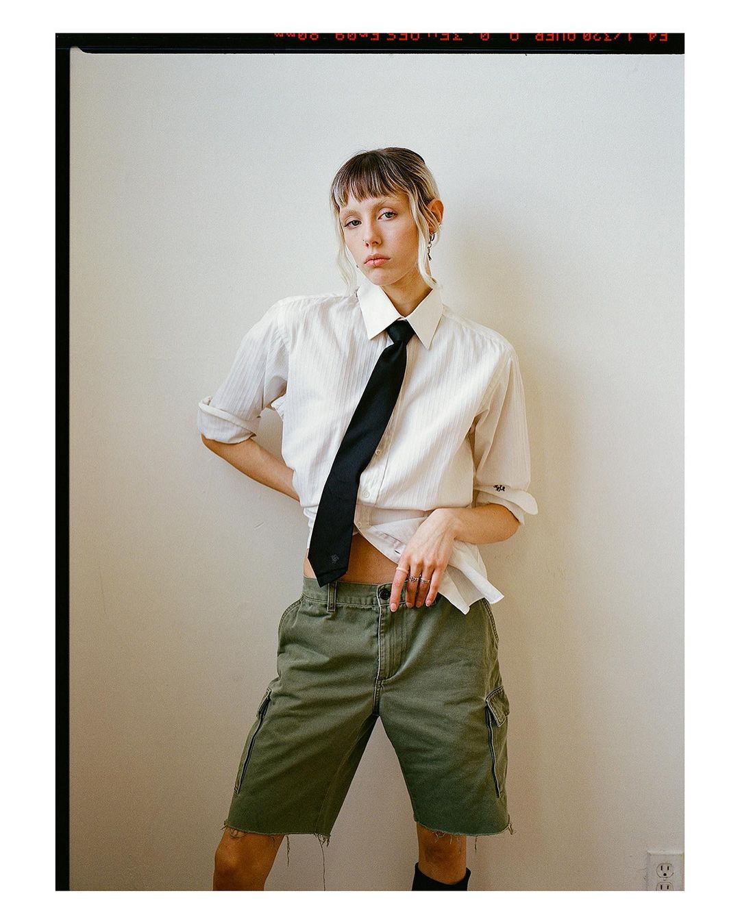Woman in unbuttoned shirt, black tie, and green cargo shorts posing against a white wall in an indoor environment with natural light.