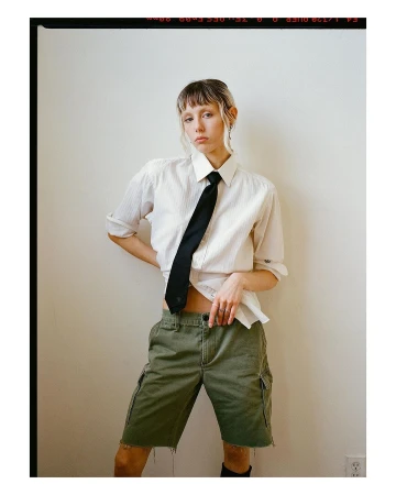 Woman in unbuttoned shirt, black tie, and green cargo shorts posing against a white wall in an indoor environment with natural light.