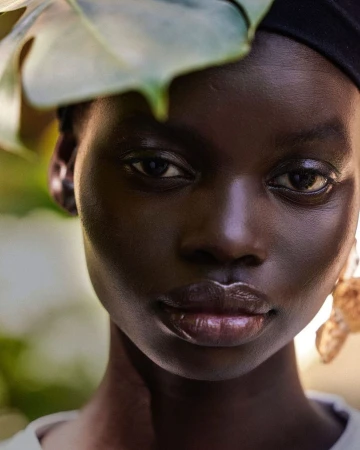 Close-up portrait of a black woman wearing a dark head wrap with a large green monstera leaf resting near her head