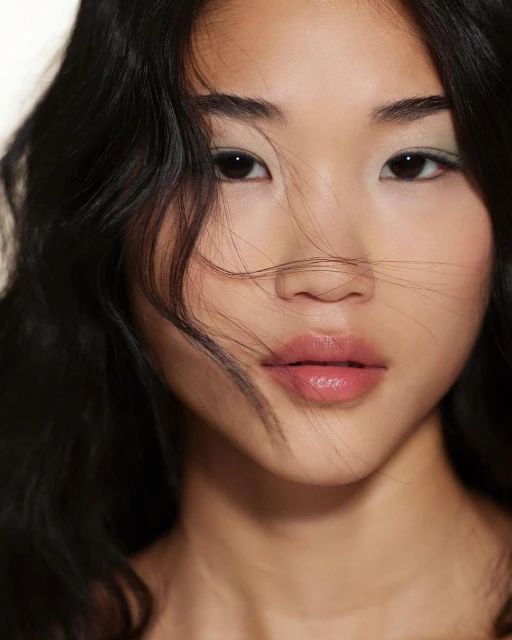 Close-up portrait of a young Asian woman with black wavy hair, light green eyeshadow, and nude lipstick against a soft white background.