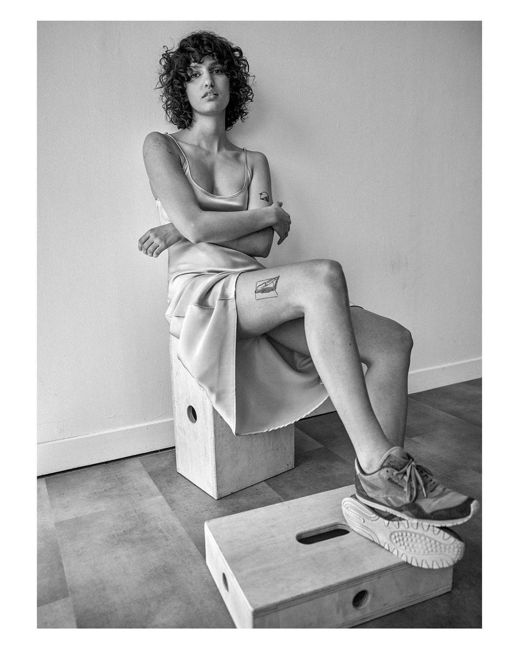 Monochrome portrait of a woman with curly hair in a satin dress sitting on a wooden box in an indoor setting, gazing at the camera.