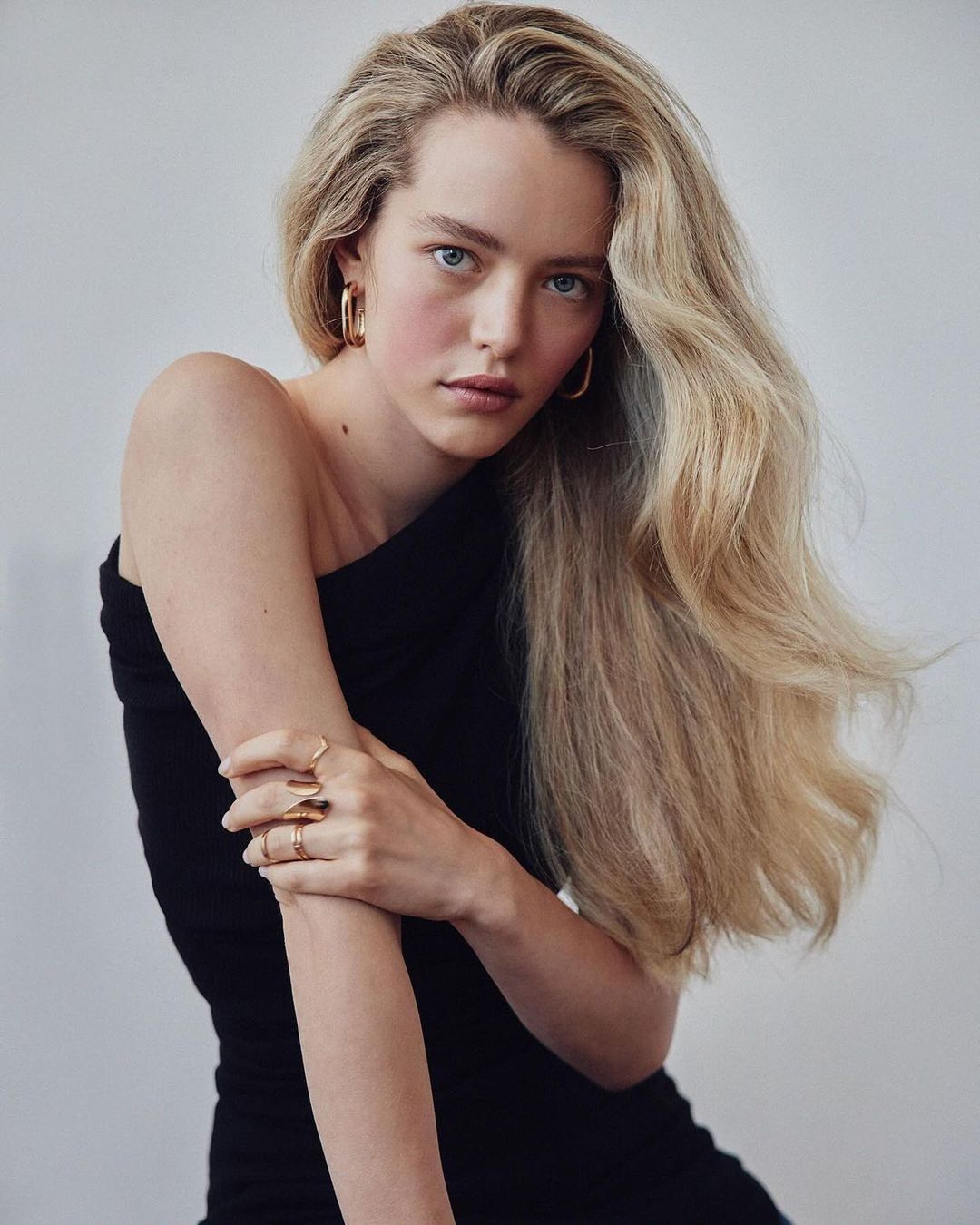 Portrait of a blonde woman in a black dress wearing gold jewelry against a white background in a studio, medium shot.