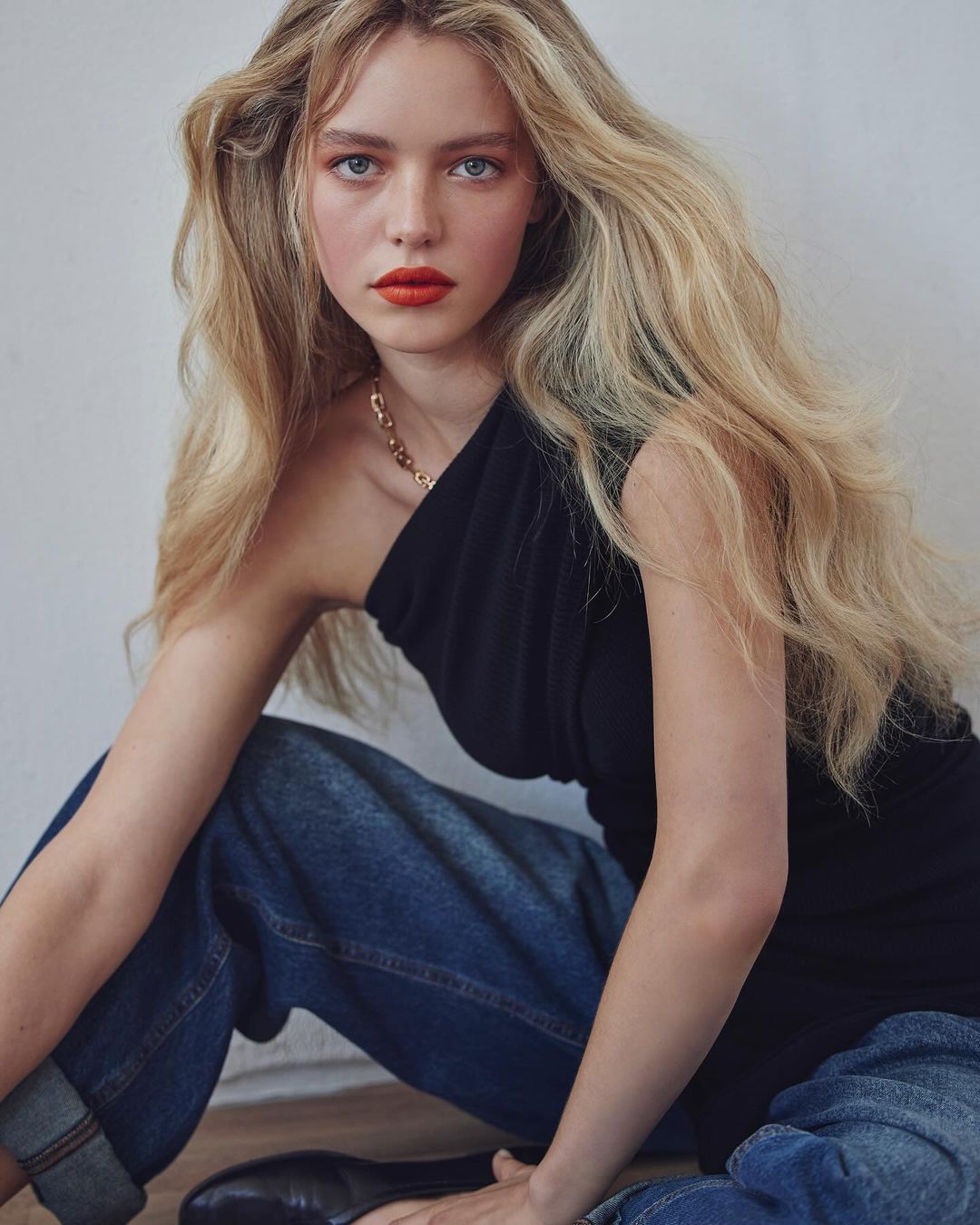 Blonde female model with bold red lips wearing a black one-shoulder top and blue jeans by Alvaro Goveia
