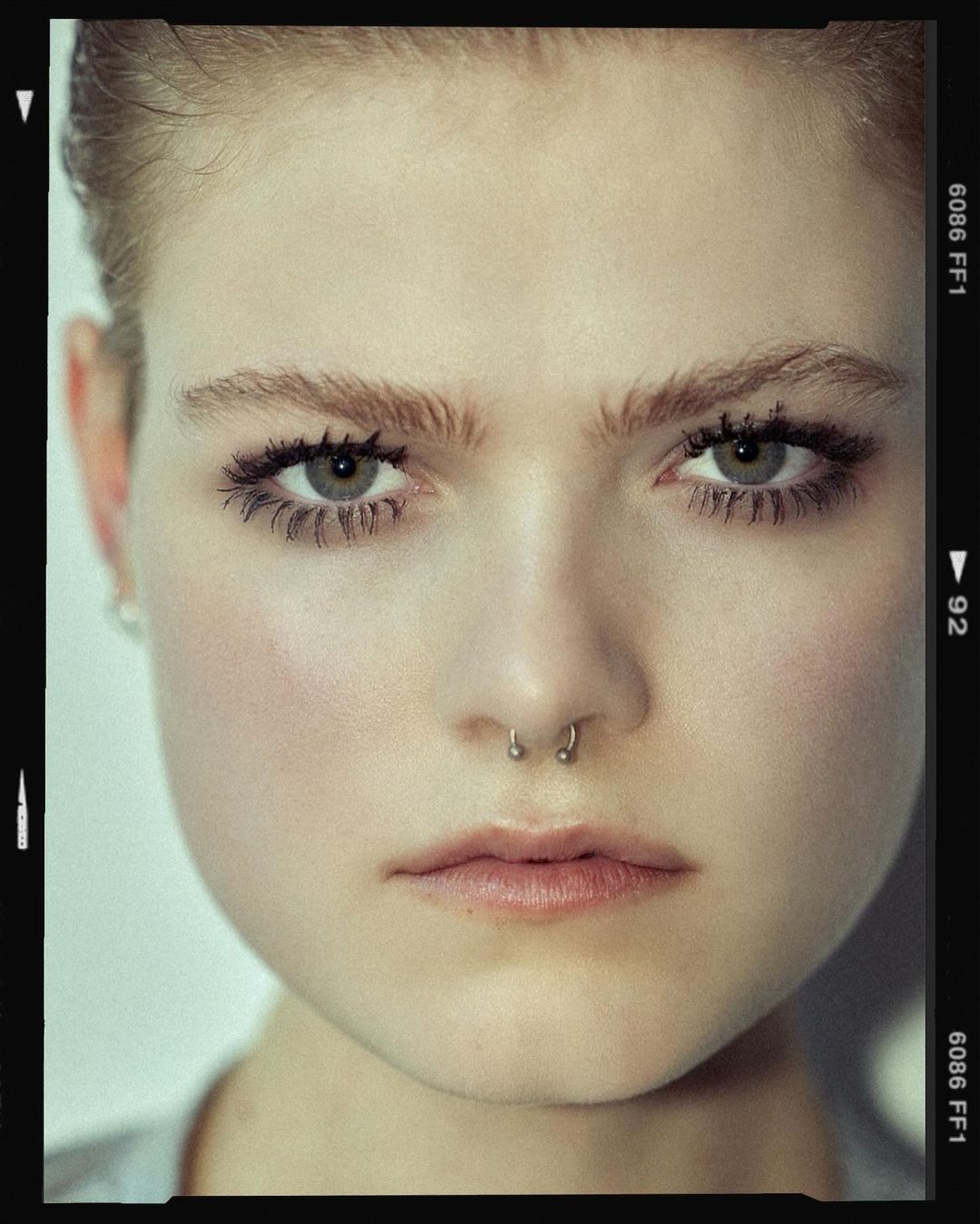 Close-up portrait of a young adult woman with light skin, light eyebrows, and minimal makeup, wearing a nose ring and gazing at the camera.