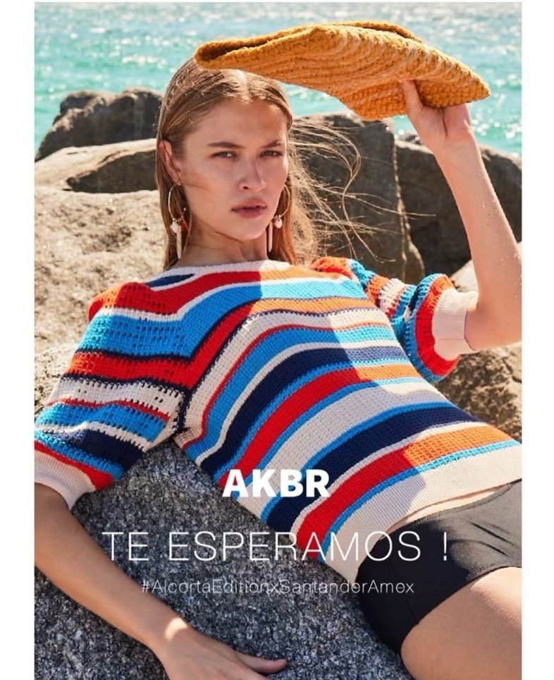 A fashion shot of a woman in a striped sweater and a straw hat on a rocky beach with the ocean in the background.