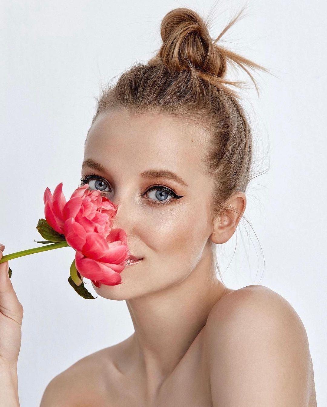 Close-up of a woman with her hair in a bun, holding a peony near her face against a white background in a beauty shot.