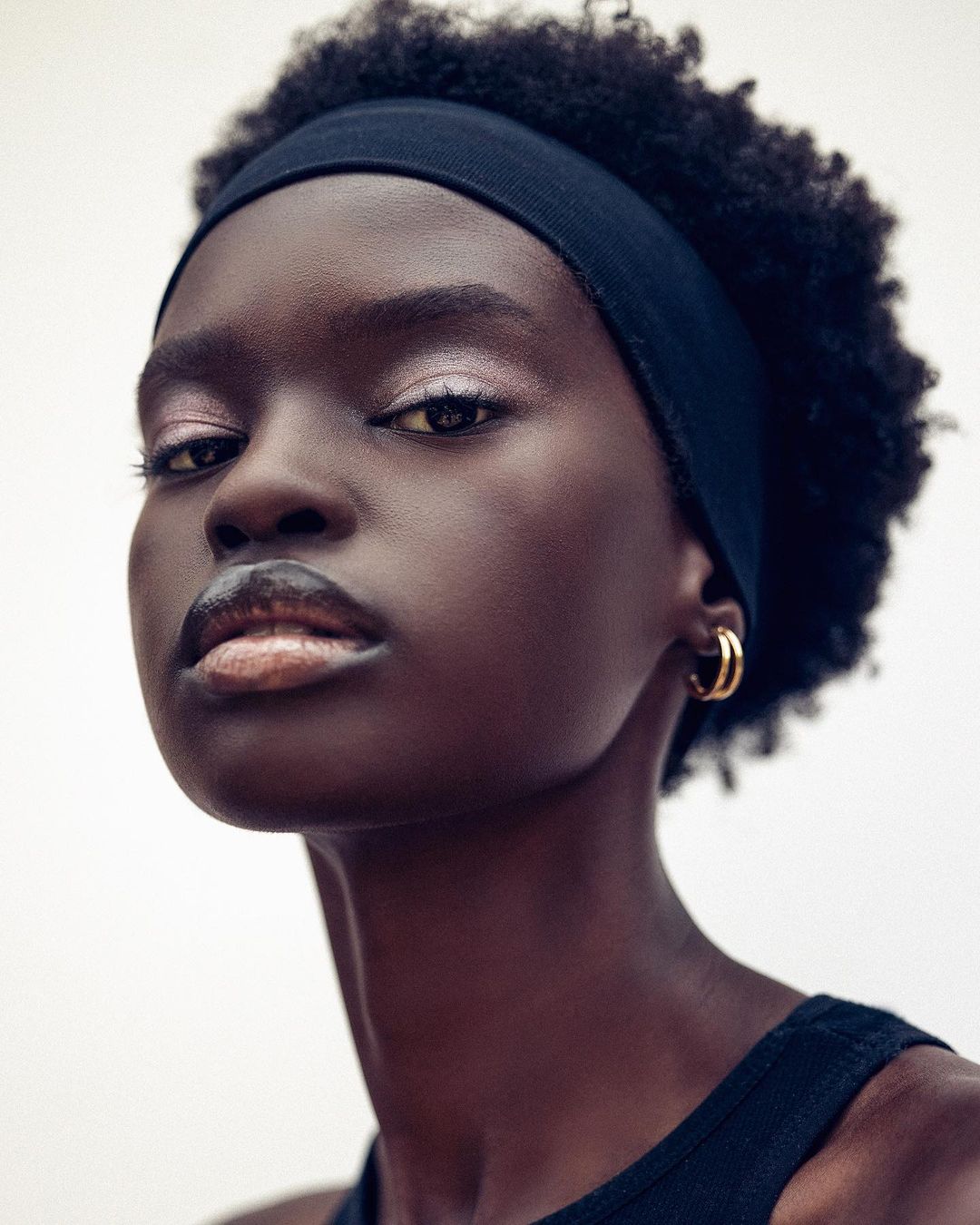 Close-up portrait of a Black woman with dark hair and a headband, wearing gold hoop earrings, against a neutral background.