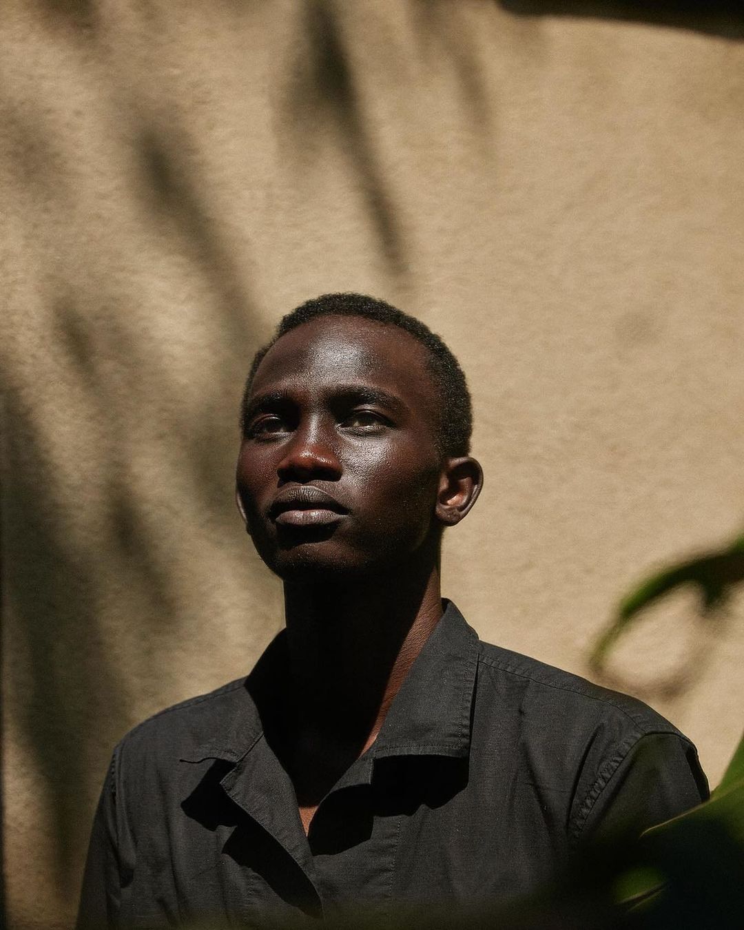 Portrait of an adult male with dark skin in black button-down shirt in partial shadow with a neutral expression looking slightly upward.