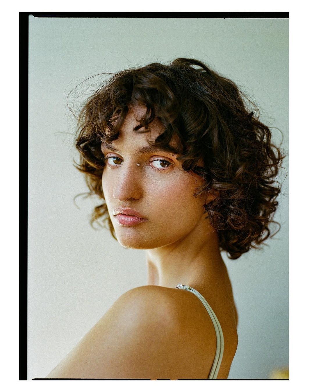 Portrait of a woman with short curly brown hair and a piercing looking over her shoulder against a plain background