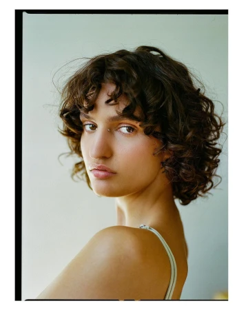 Portrait of a woman with short curly brown hair and a piercing looking over her shoulder against a plain background