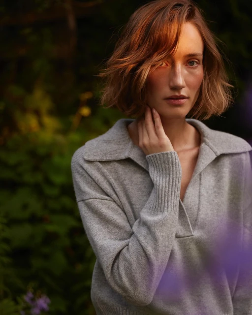 A young woman with auburn hair poses outdoors in a grey sweater, her hand near her neck, with soft lighting and lush green foliage.