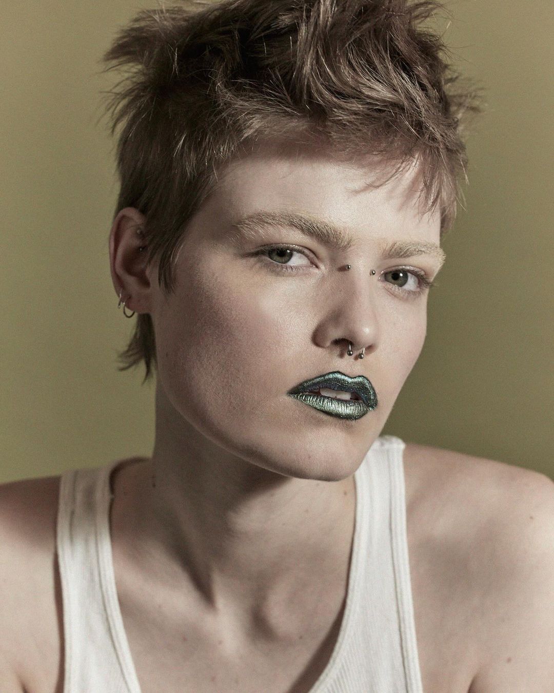Close-up portrait of an adult woman with short brown hair, fair skin, nose and eyebrow piercings, and metallic green lipstick against a tan background.