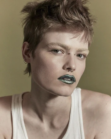 Close-up portrait of an adult woman with short brown hair, fair skin, nose and eyebrow piercings, and metallic green lipstick against a tan background.