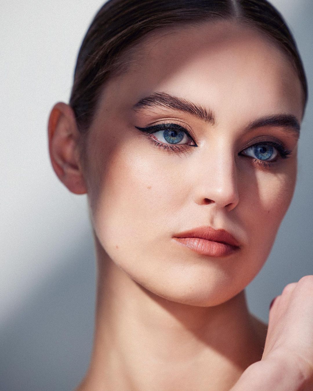Close-up portrait of a young adult woman with light skin, blue eyes, and brown hair, showcasing beauty and makeup in a brightly lit studio.