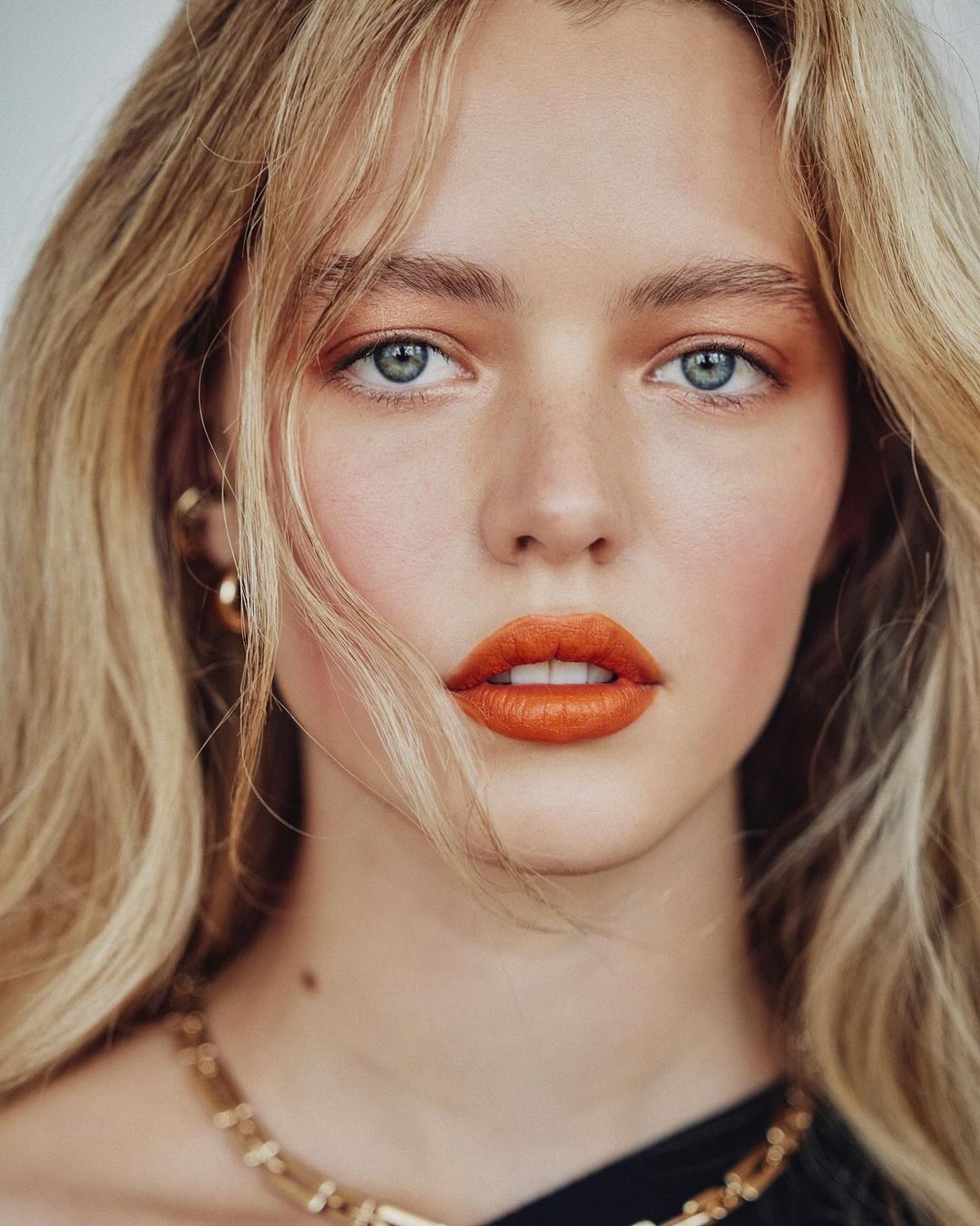 Close-up portrait of a woman with blonde hair, bright orange lipstick, blue eyes, and gold jewelry against a neutral background.
