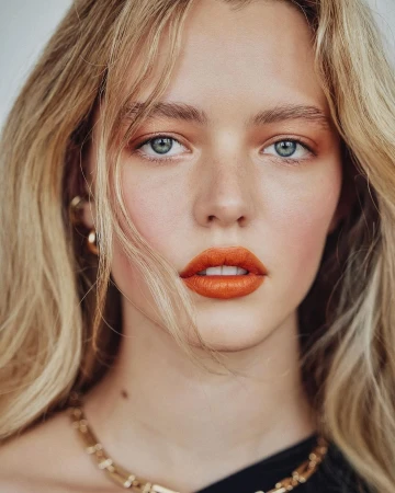 Close-up portrait of a woman with blonde hair, bright orange lipstick, blue eyes, and gold jewelry against a neutral background.