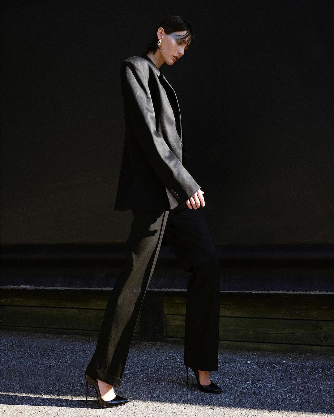 A woman in a black blazer and pants walks in front of a dark background. The lighting is bright, and she wears gold earrings.