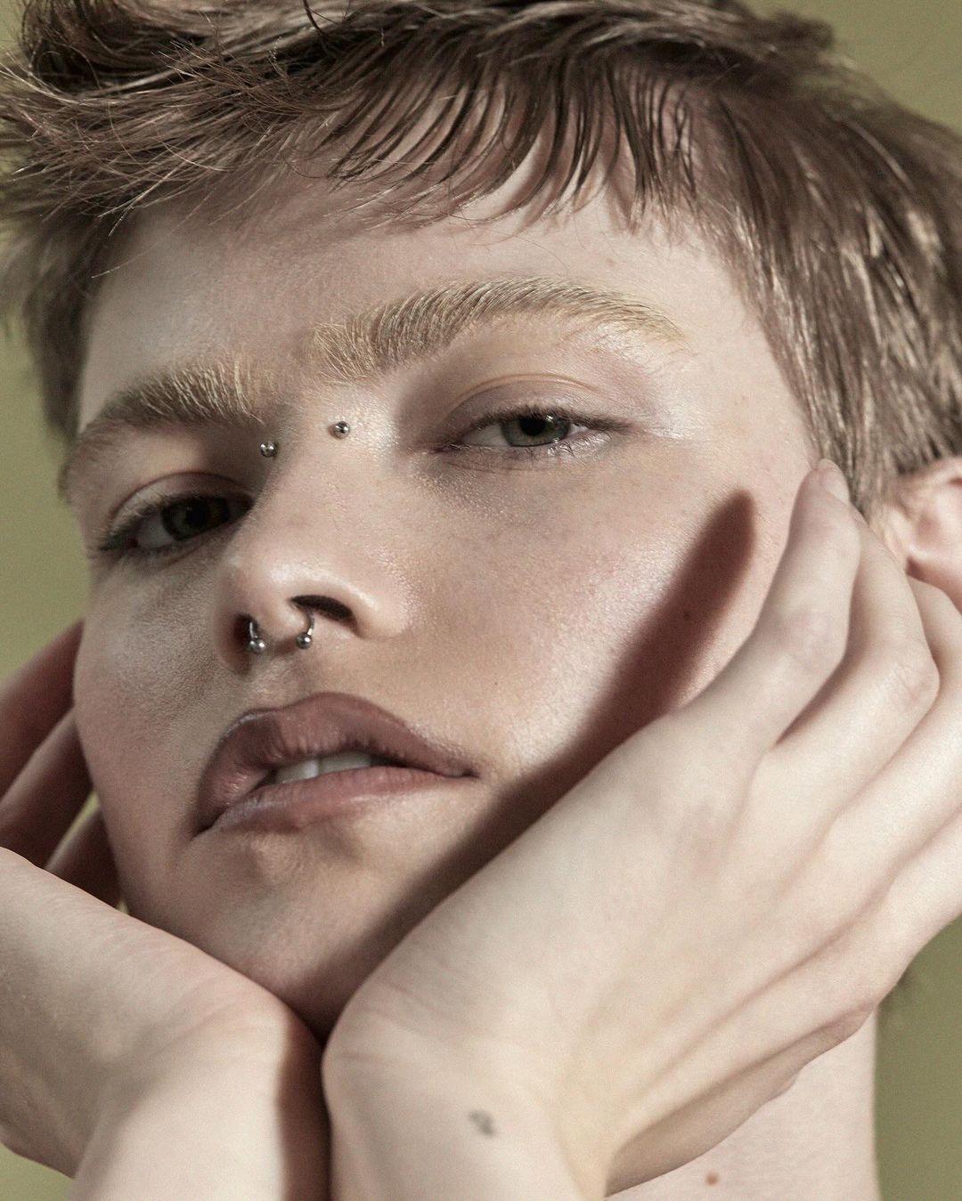 Close-up portrait of a young adult with short hair, facial piercings, and their hands framing their face against a solid olive green backdrop.