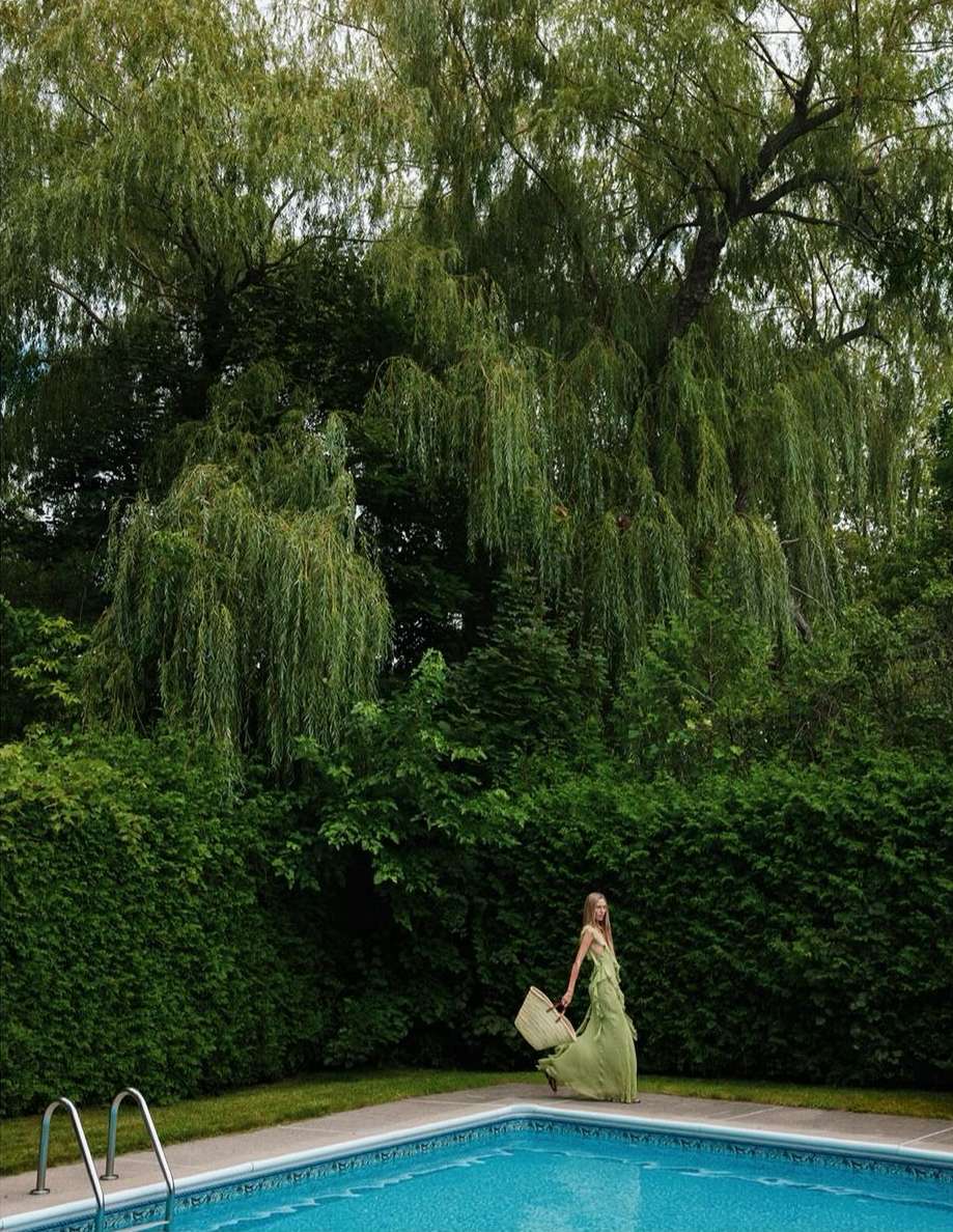 Woman in a green dress carrying a bag walks by a blue swimming pool in a backyard with a large willow tree and green foliage.