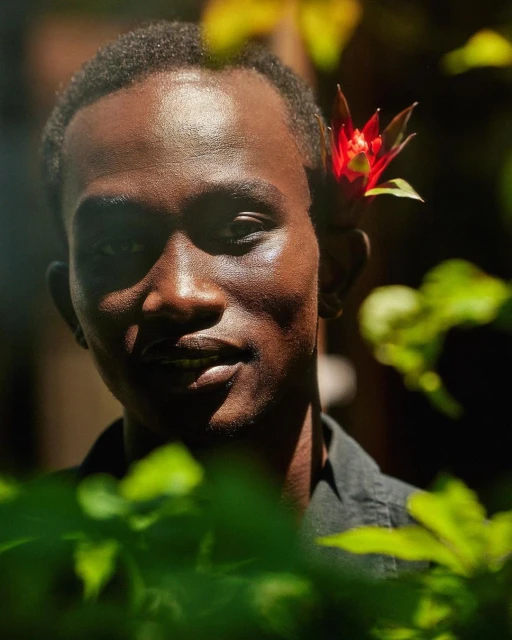 Close-up of a dark-skinned adult male with a red flower behind his ear, set against a backdrop of green foliage in natural light.