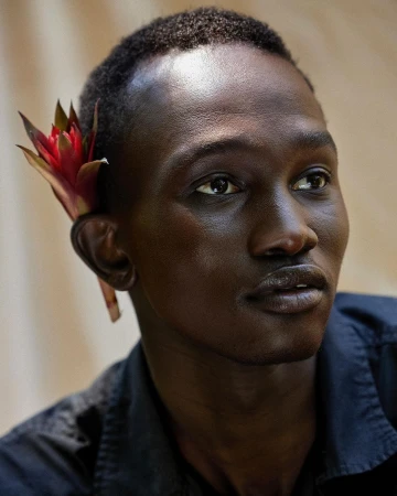 Portrait of a Black man wearing a red flower tucked behind his ear, photographed by Alvaro Goveia.
