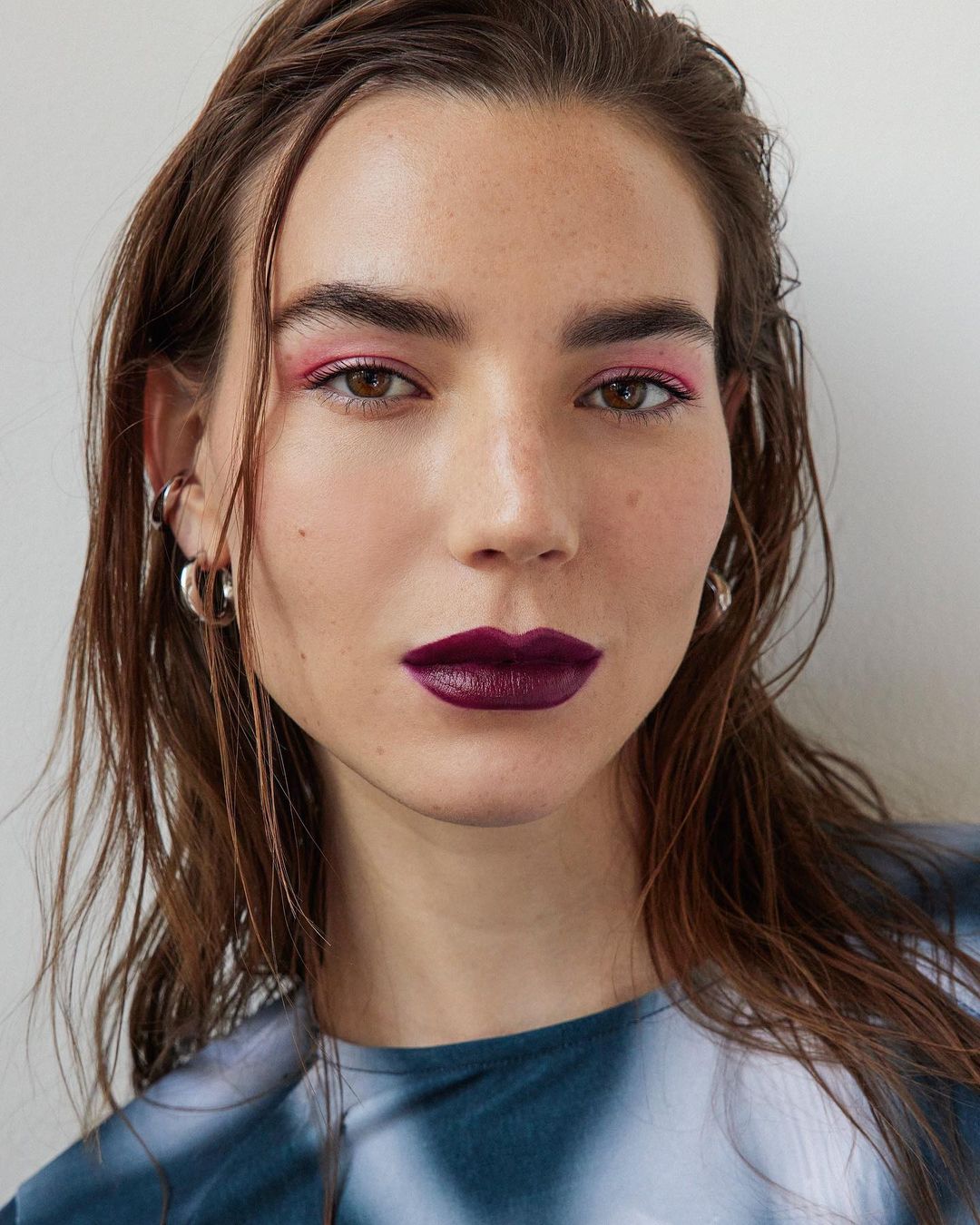 Headshot of a woman with dark hair and plum lipstick, wearing a blue tie-dye shirt against a neutral background, beauty product advertisement.