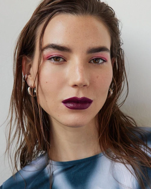 Headshot of a woman with dark hair and plum lipstick, wearing a blue tie-dye shirt against a neutral background, beauty product advertisement.