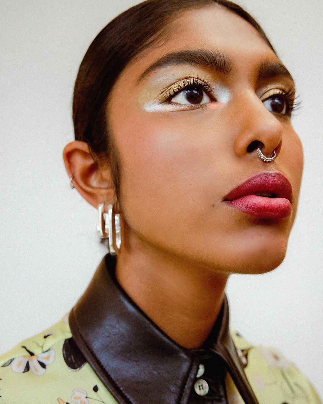Close-up portrait of a woman with colorful eyeshadow, a nose ring, and a floral shirt with a dark leather collar, in a brightly lit studio.