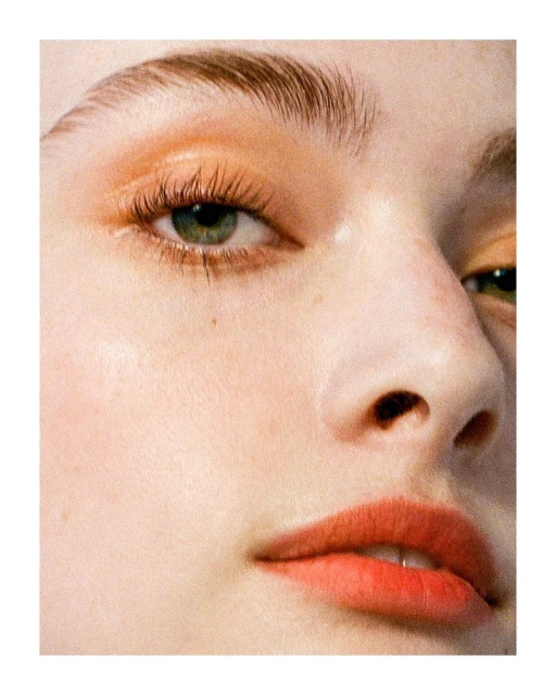 Close-up beauty shot of a young adult woman's face with dramatic peach eyeshadow, thick eyebrows, and coral lipstick.