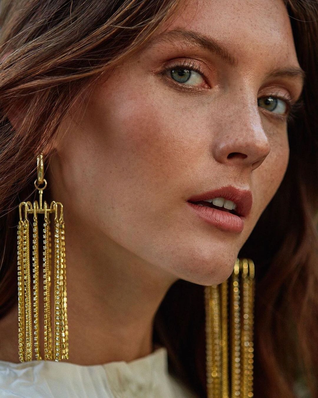 Close-up portrait of a fair-skinned adult woman with reddish-brown hair wearing dangling gold earrings with a neutral expression.