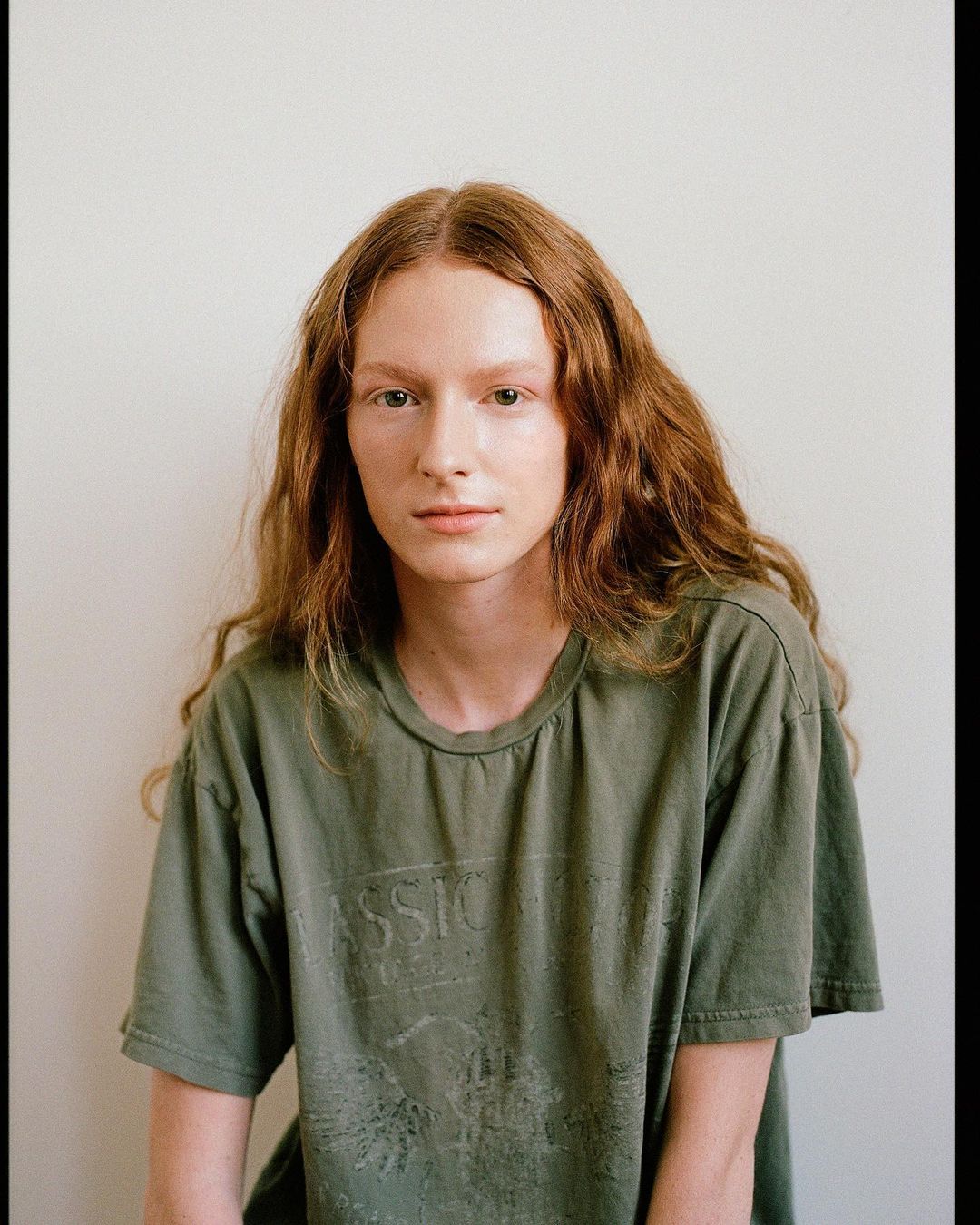 Portrait of a young adult female with long auburn wavy hair wearing an olive green t-shirt, set against a plain white backdrop.