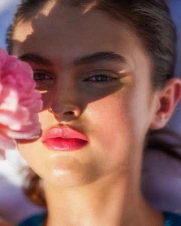 Close-up portrait of a woman with vibrant pink lips next to a pink flower, photographed by Alvaro Goveia.