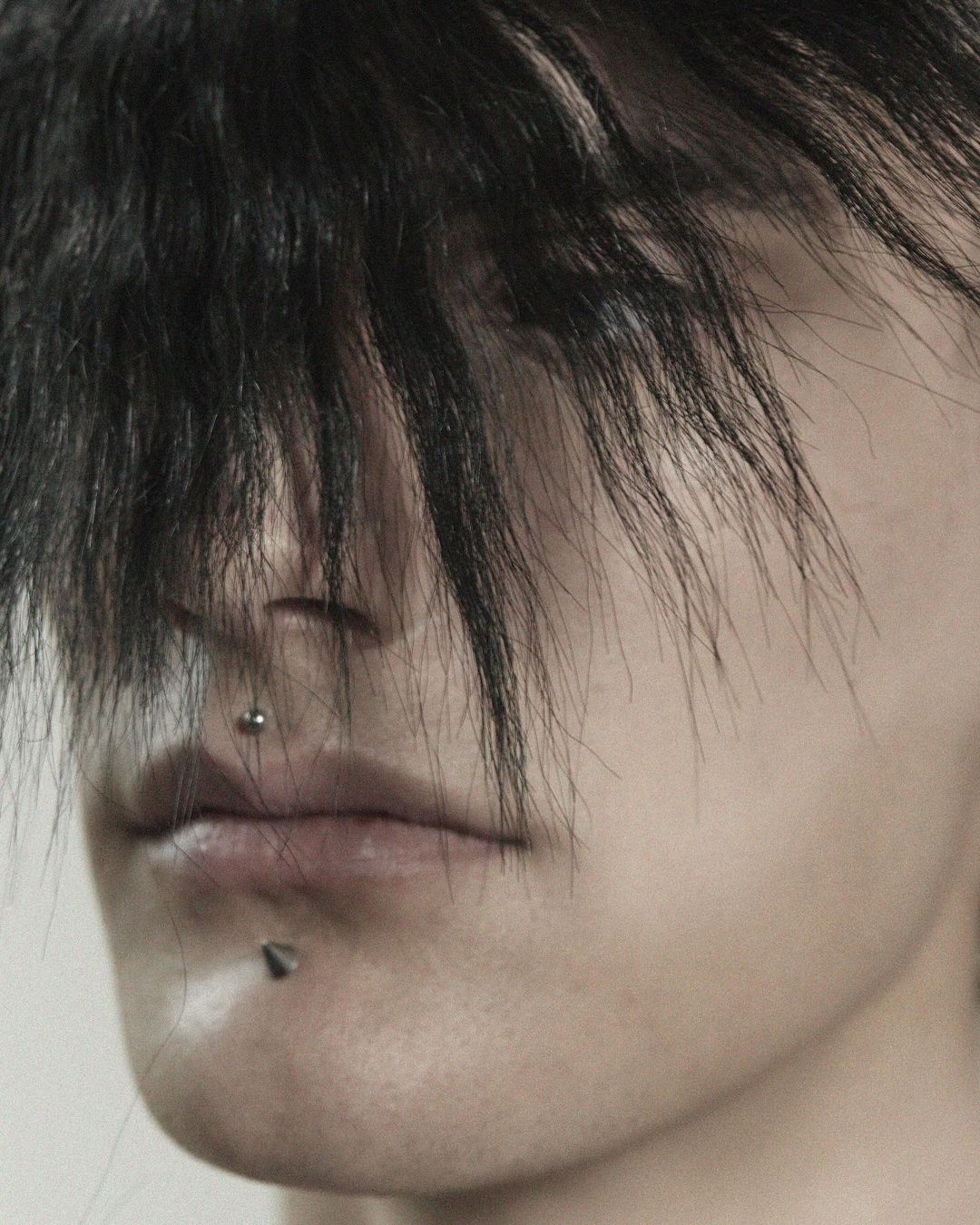 Close-up portrait of a person with dark hair covering their eyes and lip and nose piercings, set against a neutral background.