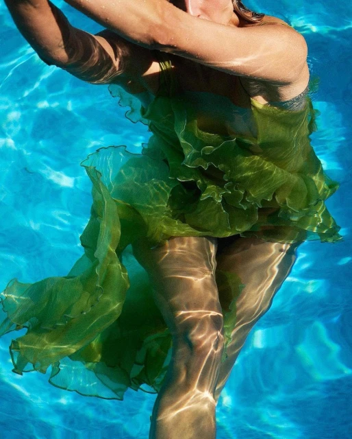 Person in a green dress swimming underwater in a bright blue swimming pool, with sunlight creating patterns on the skin and dress.
